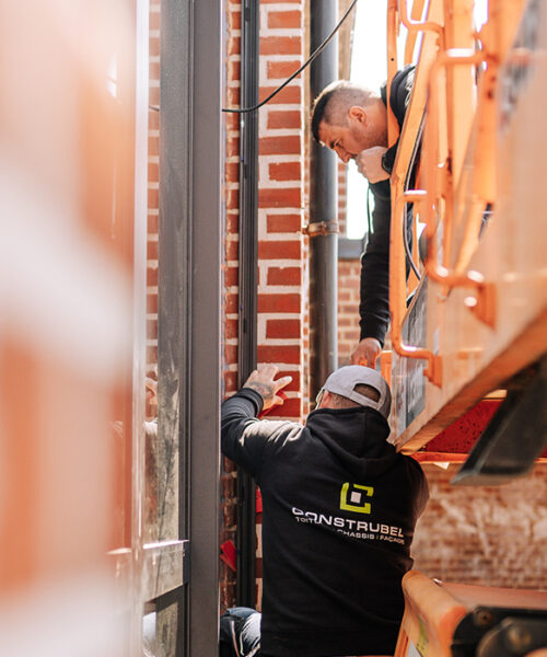 Valeur - Esprit d'équipe & collaboration Deux ouvriers qui travaillent à la pose de châssis sur un bâtiment public en briques